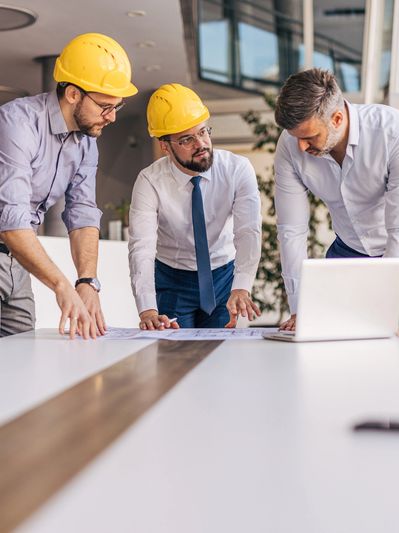Three professionals in hard hats reviewing blueprints around a table with a laptop.