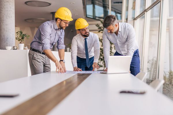 Three engineers in helmets reviewing plans at a conference table.