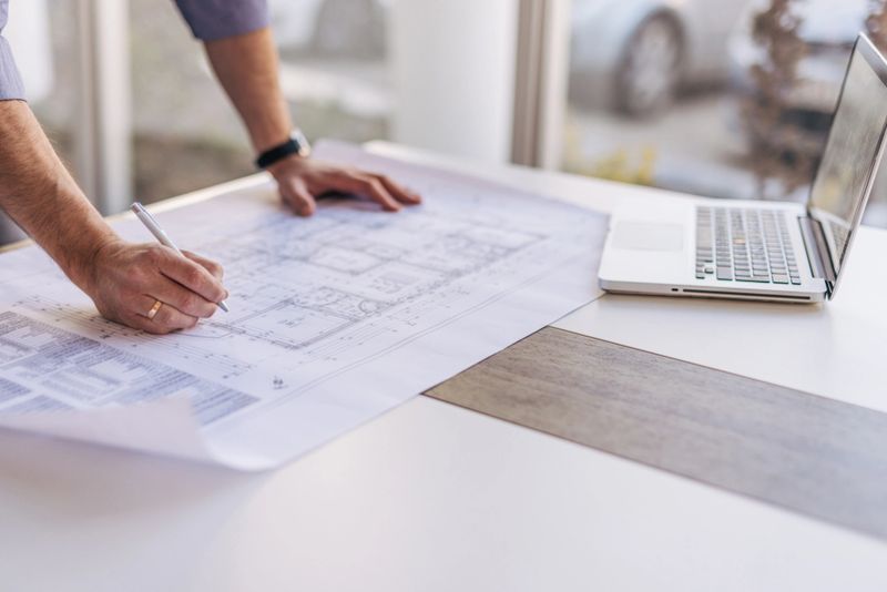 Architect reviewing blueprints and making corrections with a pen, while a laptop computer sits nearby on the desk. Focusing on detailed plans for construction projects