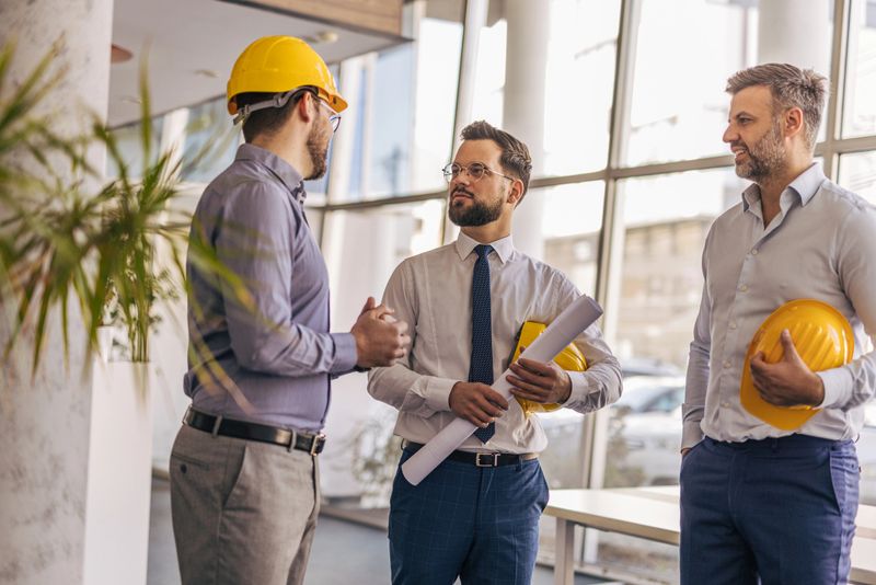 Three male engineers and architects discussing a new project in a modern office, collaborating over blueprints while wearing hardhats and dressed in formal attire