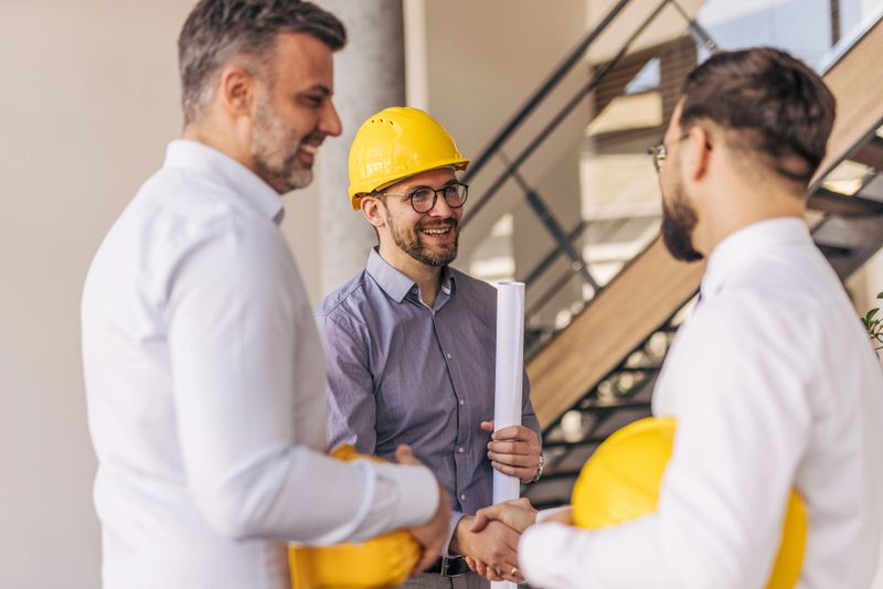 Three architects shaking hands on a construction site after finalizing an agreement, one holding blueprints, embodying teamwork and collaboration in a successful project