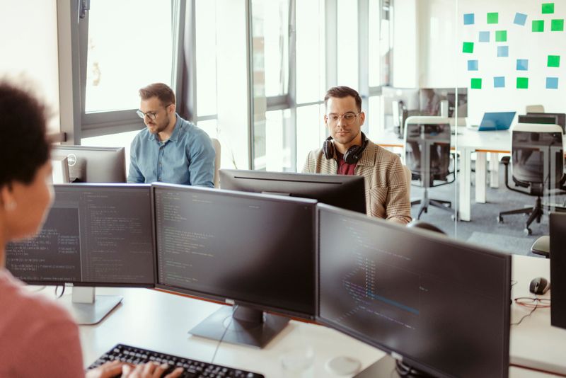 In a bright, open-plan office, a group of programmers works together, surrounded by dual monitors