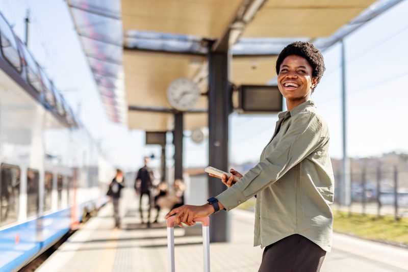 A cheerful woman with luggage smiles while standing at a sunny train station platform, holding her phone and ready for travel. The scene conveys excitement, anticipation, and a positive traveling experience.