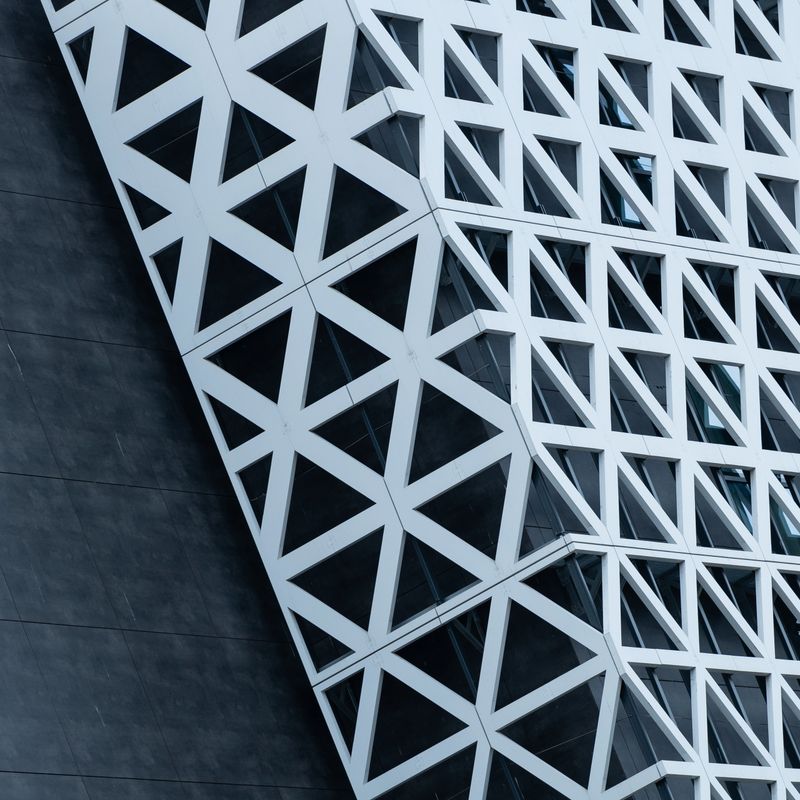 This captivating image showcases a modern business building adorned with a sleek glass facade with metal triangles, standing proudly in the enchanting daylight blue light. The gentle, cool hues of the sky cast a soft and serene glow upon the structure, creating an atmosphere of tranquility and sophistication. The building's reflective glass exterior mirrors the surrounding sky environment, creating a mesmerizing interplay of light and reflections. This image serves as a stunning abstract background, exuding a sense of contemporary elegance and professional allure, making it ideal for a wide range of design and business applications. Copy space.