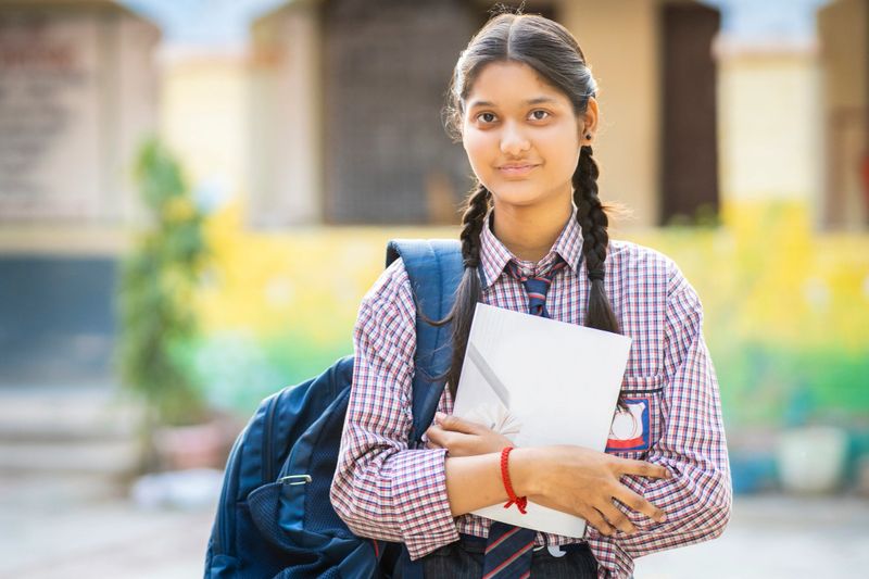 Portrait of happy indian teenage girl student in school uniform holding a book in hand standing in the school yard and looking at the camera with confident smile.