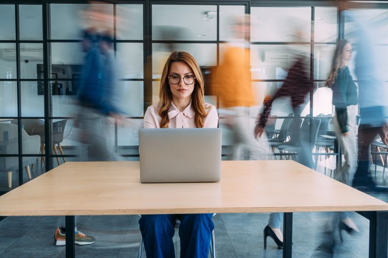 Shot of businesswoman sitting on desk in her office and using laptop. Blurred motion of energetic businesspeople on the move and businesswoman working on laptop in modern office. Creative business person using laptop in the workplace while her colleagues are walking in blurred motion. Busy hours in a board room during working hours while contemporary businesswoman is looking at laptop and her colleagues are passing behind her in a conference room during business hours. Beautiful smiling businesswoman sitting on desk in a busy office, coworkers walk behind her in blurred motion. Shot of a female creative with her colleagues blurred in the background. Woman working at a creative office using her computer and people moving at the background.