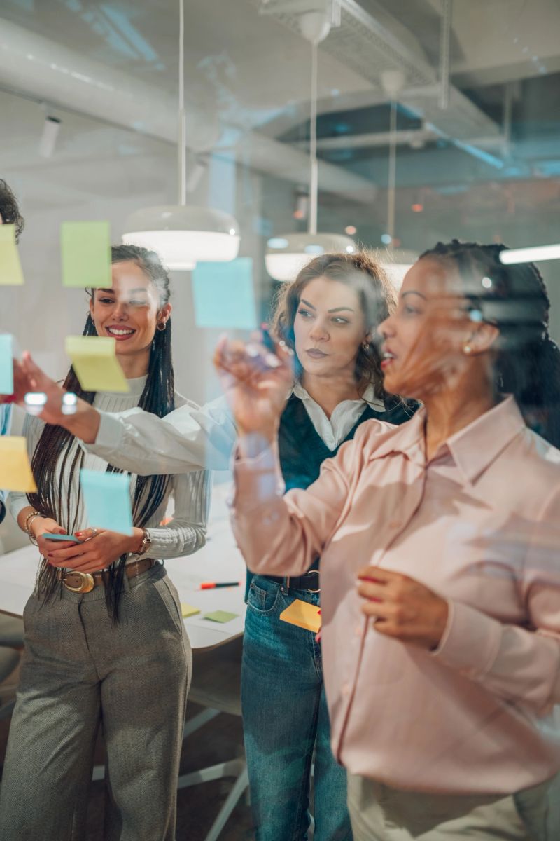 Four businesswomen are using colorful sticky notes on a glass wall, brainstorming and planning a new strategy