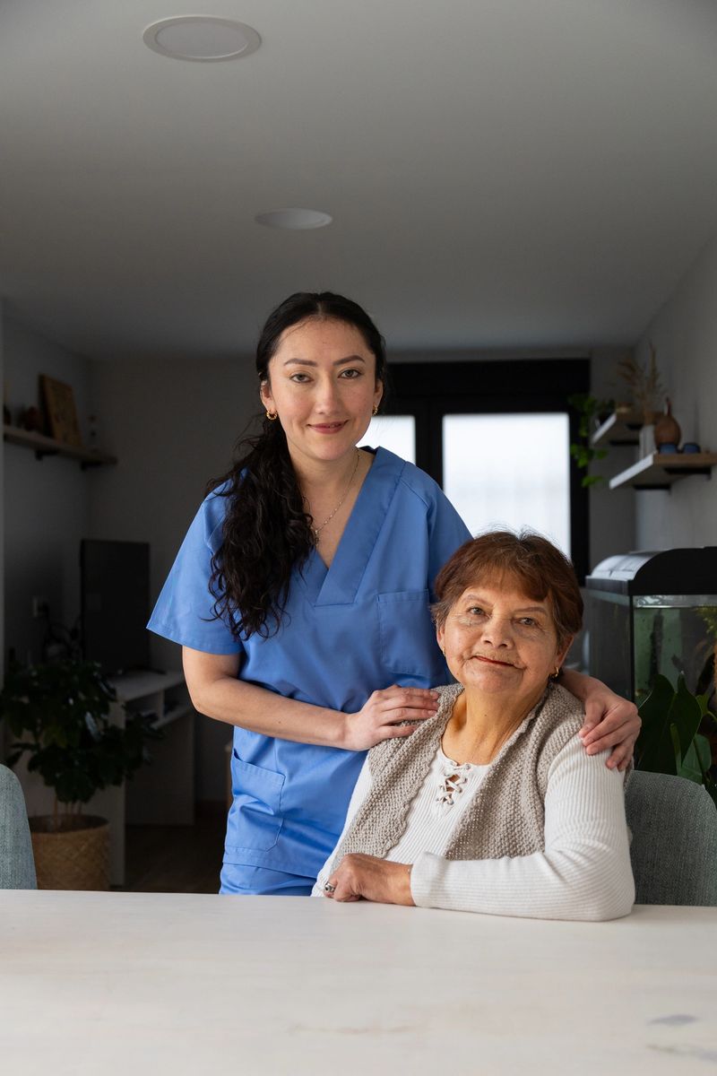 Young nurse supporting an elderly woman seated at a table in a cozy home environment, providing compassionate care and assistance