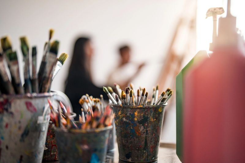 Close-up of colorful paintbrushes in jars in a creative studio, with blurred diverse students painting in the background.