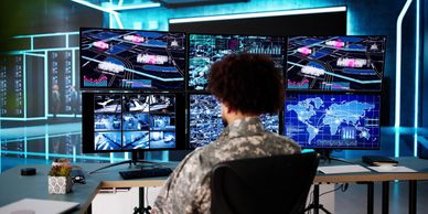 A soldier monitors multiple surveillance and data screens in a high-tech control room.