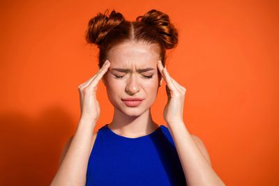 Young woman with headache holding temples against orange background.
