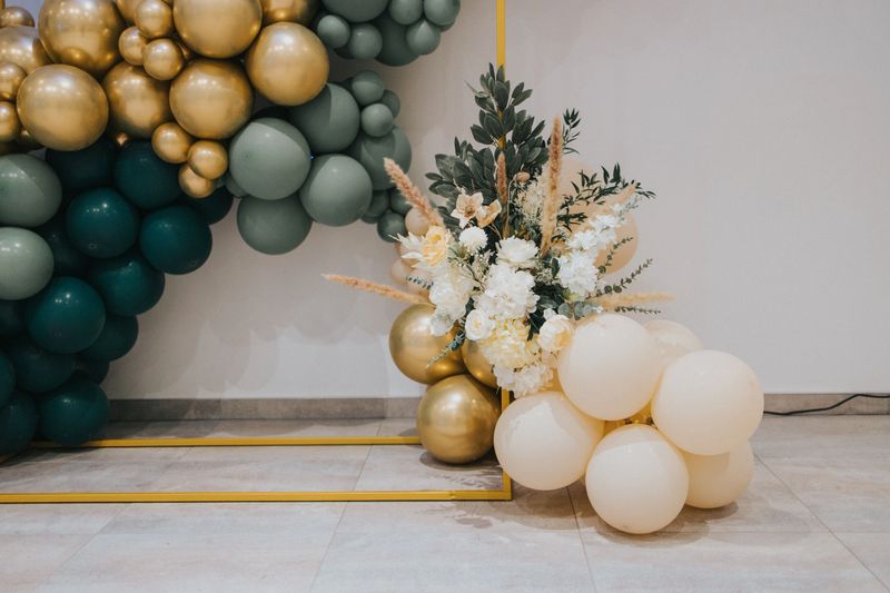Elegant balloon arch with gold, green, and white balloons, complemented by floral arrangements, creating a sophisticated event decor.