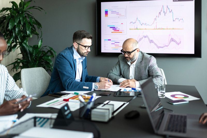 Two businesspeople collaborate in an office setting, analyzing business charts and documents on a table, while digital graphs display on a screen, showing teamwork and analysis in a professional environment.