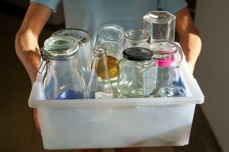 A person holding a plastic container filled with empty glass jars and bottles, preparing for waste sorting and recycling as part of a sustainable lifestyle and environmental conservation effort.