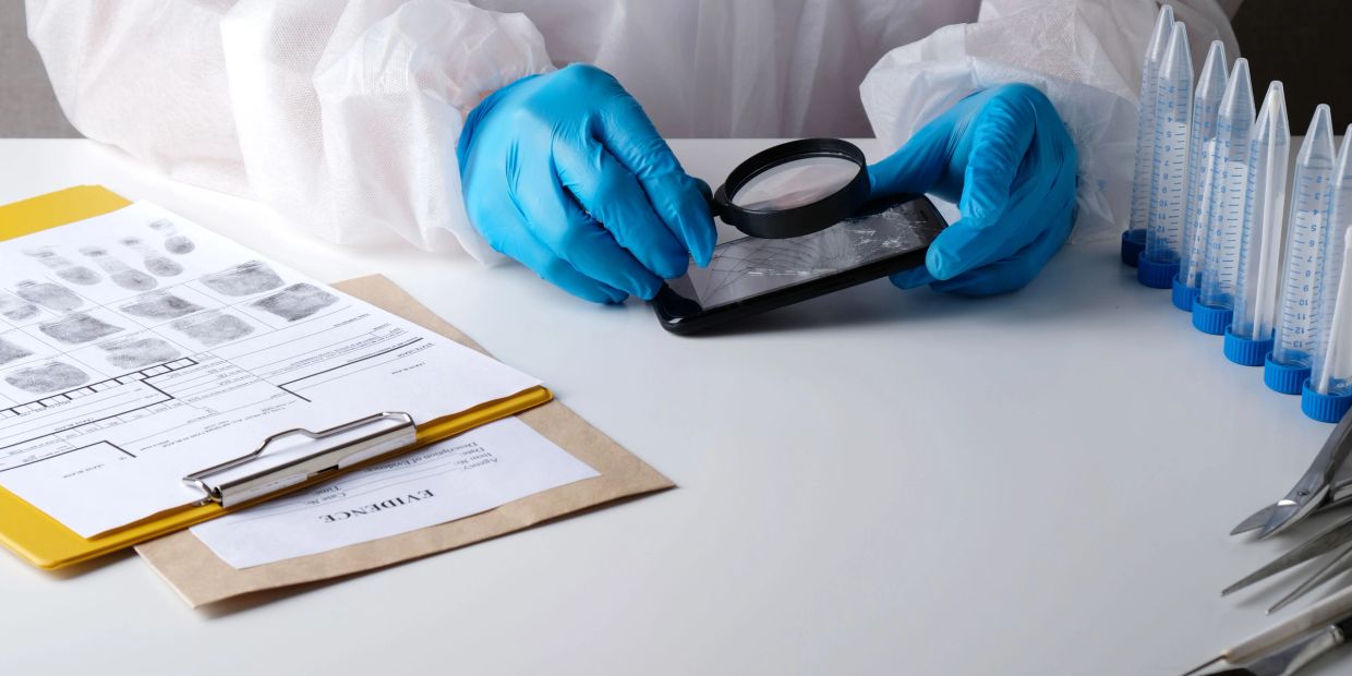 Forensic expert examines a cracked phone with a magnifying glass in a lab setting.