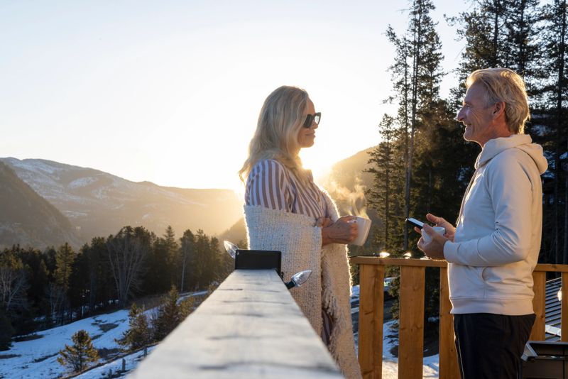 Mature couple chat on chalet deck at sunrise in winter, with phone and hot drink, Canadian Rockies