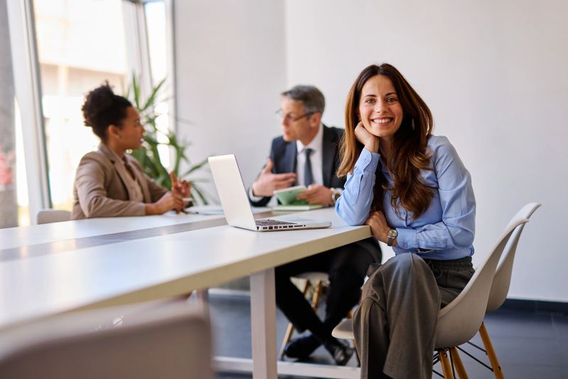 Businesswoman is smiling during a meeting with colleagues in a modern office, she is sitting at a table with a laptop and looking at the camera