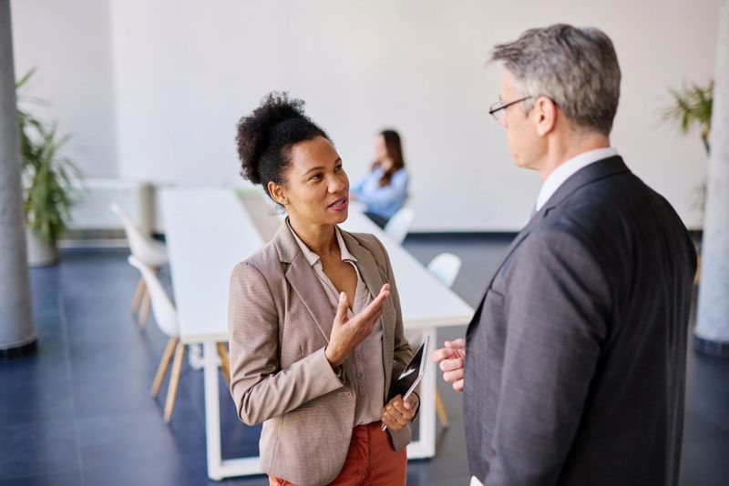 Businesswoman holding a tablet while explaining strategy to a businessman during a meeting in a modern office, with colleagues collaborating in the background