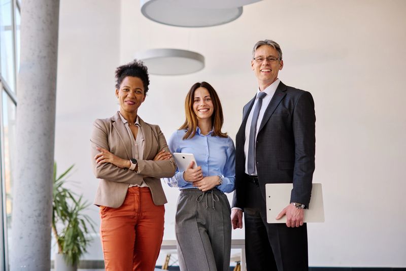 Three business people, a woman with crossed arms, a woman holding a digital tablet and a man with glasses holding a clipboard, are posing for a portrait in a modern office, smiling at the camera