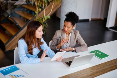 Two women discussing work at a desk with a laptop and tablet.