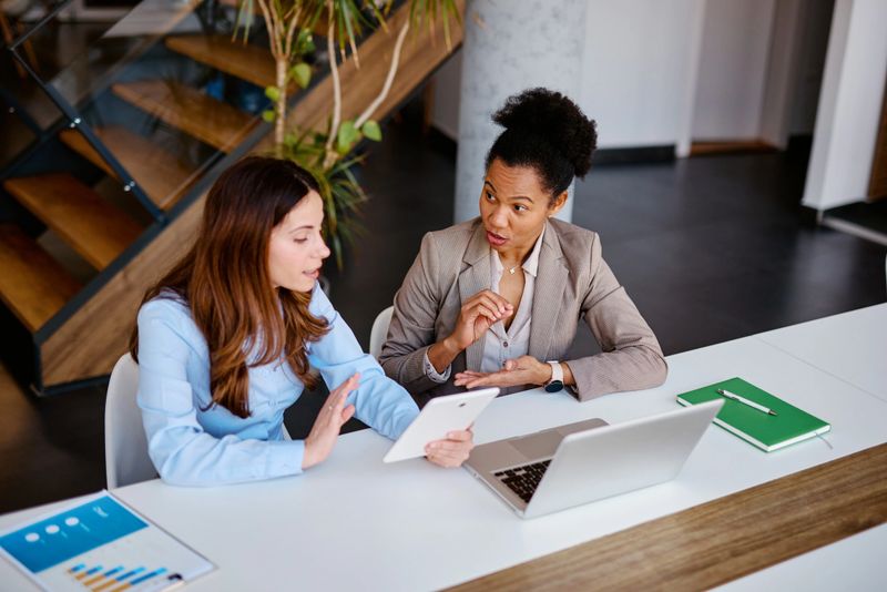 Two businesswomen are sitting at a white table in a modern office, using a digital tablet and discussing work, with a laptop and notepad nearby