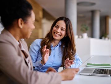 Two women smiling and discussing work at a laptop in an office.