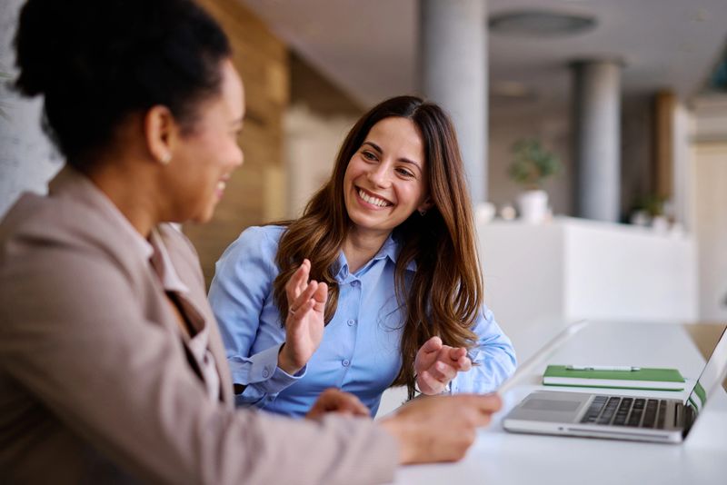Two smiling businesswomen discussing a project in a modern office, collaborating with a laptop and documents, exemplifying teamwork and effective communication in a professional setting
