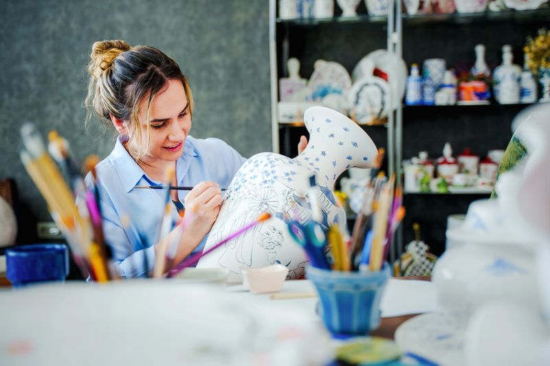 In a ceramics workshop, a woman is drawing hand-painted designs on a large vase. she is painting  carefully. The workshop environment includes ceramic products and painting materials. A scene reflecting the themes of art, craft, handicraft, ceramic painting, women's solidarity and creativity.