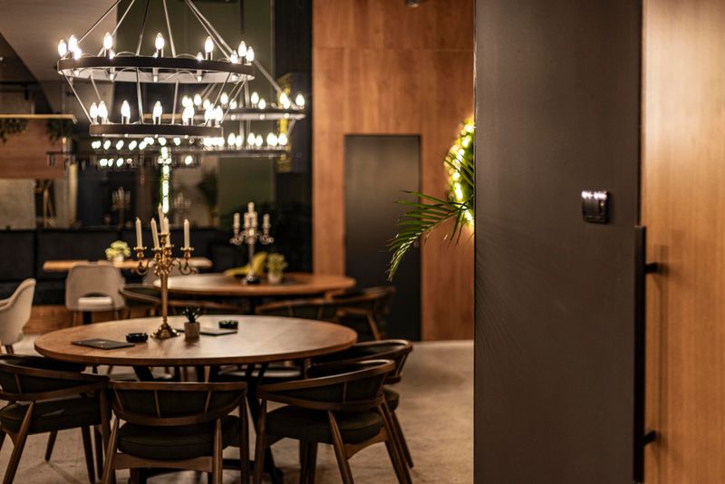 A warm and inviting modern restaurant features elegant wooden tables, stylish chairs, and soft lighting. The atmosphere is enhanced by lush greenery and decorative elements, creating a sophisticated dining experience.