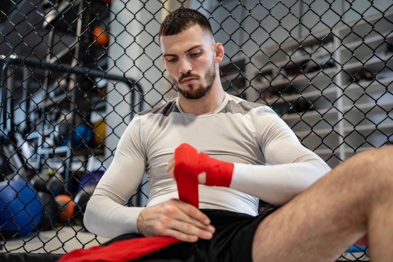A determined man wearing athletic clothing wraps his hands while sitting beside a cage in a gym. The image depicts focus and commitment to physical training and preparation for combat sports or fitness activities.