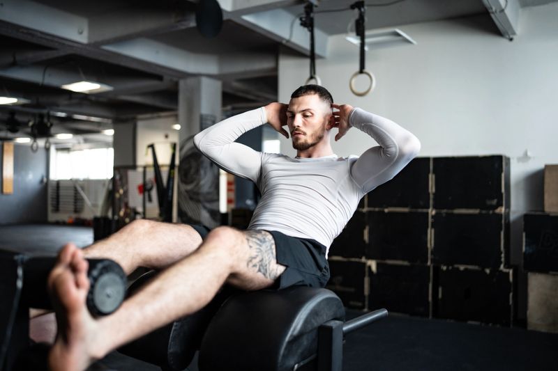 Athletic man in a gym performing a crunch exercise on a bench, surrounded by fitness equipment.