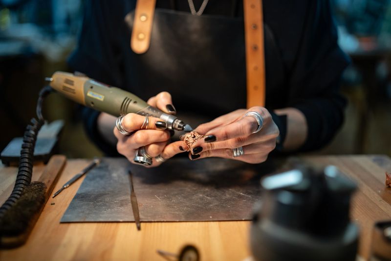 A craftswoman meticulously works on a detailed jewelry piece using a rotary tool on a sturdy workbench.
