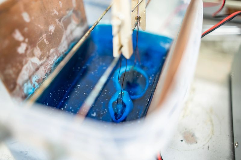 Close-up view of a metallic object undergoing electroplating in a blue solution, showing industrial precision and surface treatment techniques, highlighting technology and craftsmanship.
