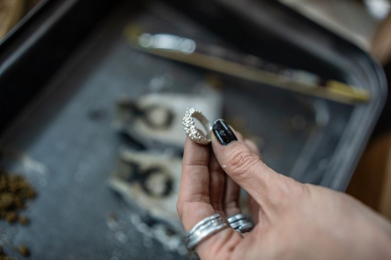 A closeup of a hand with black painted fingertips holding a delicately crafted silver ring in a professional jewelry-making workshop setting that conveys artistry, craftsmanship, and elegance.