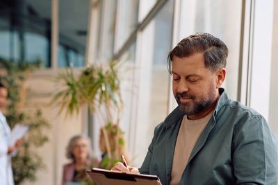 Man filling out a form in a medical waiting room with a doctor and patients in the background.