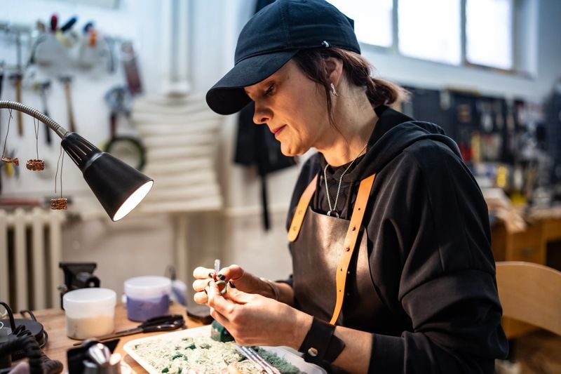 A skilled craftsperson wearing an apron and cap focuses intently on delicate jewelry work at a well-equipped workshop desk, showcasing dedication, craftsmanship, and a passion for handmade artistry.