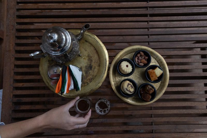 The photo features a traditional Moroccan welcome tea set, with a steaming glass of mint tea placed on an ornate silver tray. The vibrant green of the tea is topped with fresh mint leaves, and the glass is decorated with intricate patterns, reflecting the elegance of Moroccan craftsmanship. Alongside the tea, several small plates are filled with an array of colorful, sweet Moroccan pastries. Delicate cookies dusted with powdered sugar, honey-soaked treats, and flaky baklava add a touch of sweetness to the scene. The tray is adorned with a beautifully designed teapot and small serving cups, creating a warm and inviting atmosphere that embodies the Moroccan tradition of hospitality.