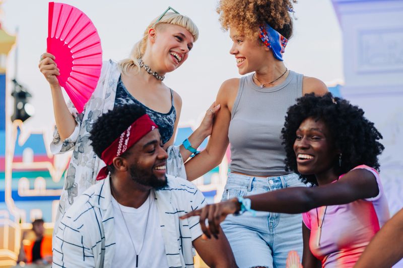 Four happy multi ethnic young adults enjoying time together at outdoor summer music festival, woman holding pink fan, colorful stage in background