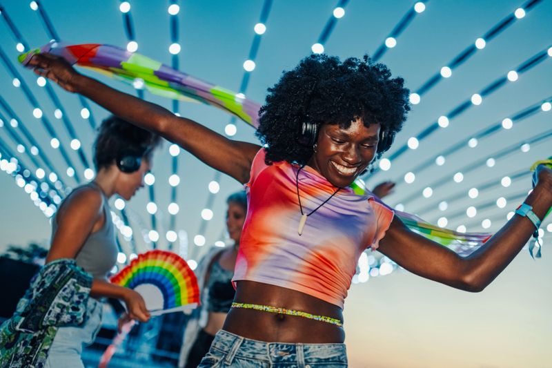 Happy young woman dancing with friends at an outdoor music festival, wearing headphones and a colorful scarf, enjoying the vibrant atmosphere under twinkling string lights
