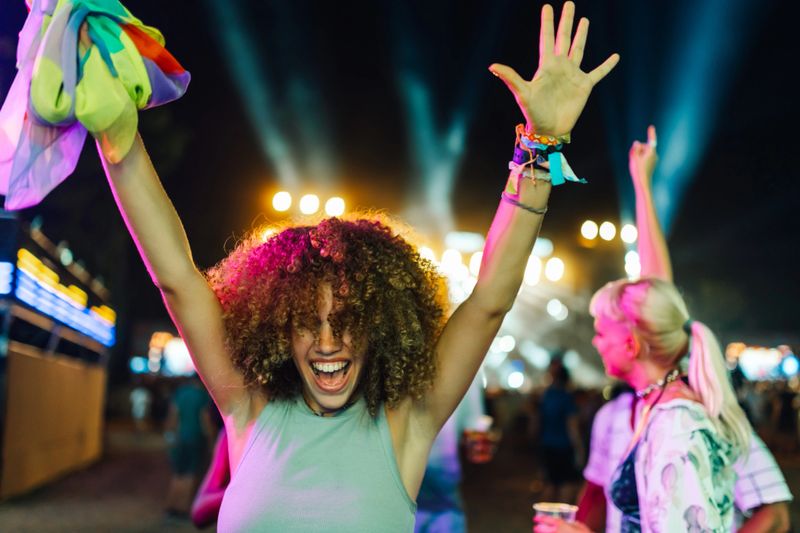 Young woman with curly hair enjoys live music, waving a rainbow scarf and dancing with raised arms at an outdoor music festival at night, expressing joy and freedom