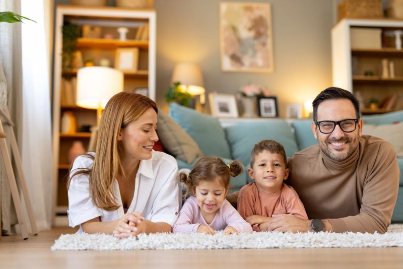 Parents and two children, a boy and a girl, lying together on a plush white rug in a living room, sharing smiles and pleasant family moments with each other.