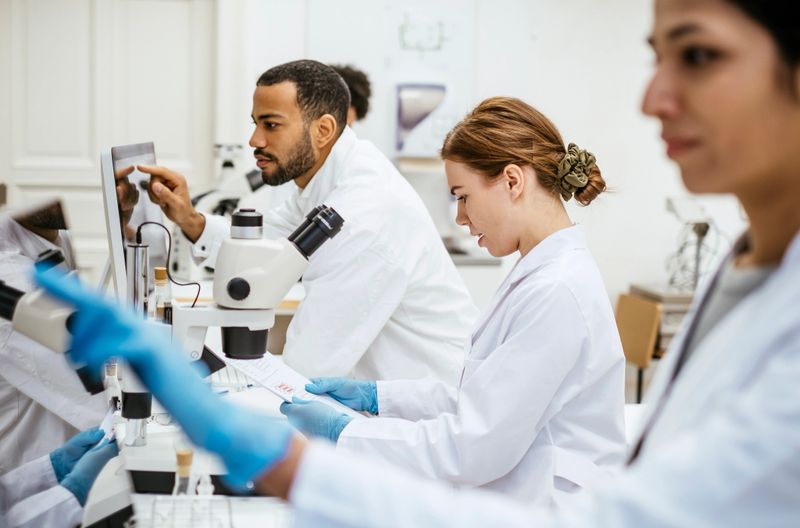 Diverse group of STEM students, including men and women, working in a university laboratory, wearing white lab coats and gloves, using microscopes and scientific tools for research and experiments.