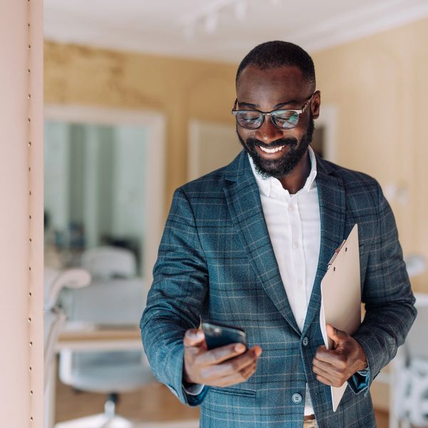 Smiling man in a suit looking at his phone while holding a clipboard.