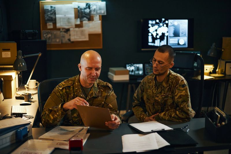 Two military personnel in uniform analyzing documents and charts in command center filled with equipment and technology. Collaboration and planning evident in their focused expressions