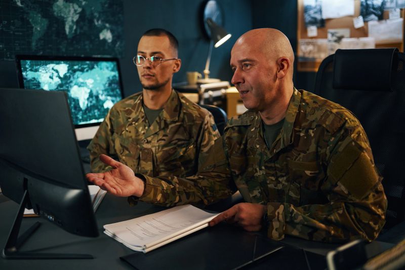 Two military personnel in fatigues examining screen and documents in strategy room with maps on wall around. Engaged in detailed strategic planning without distractions