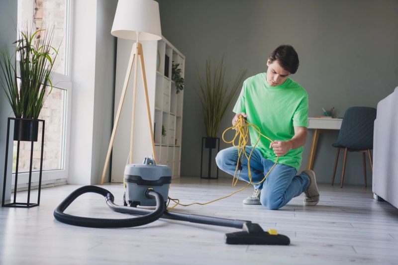 A young man in a casual t-shirt organizing a vacuum cleaner cable in a modern and well-lit living room, showcasing a homey and relaxed atmosphere.