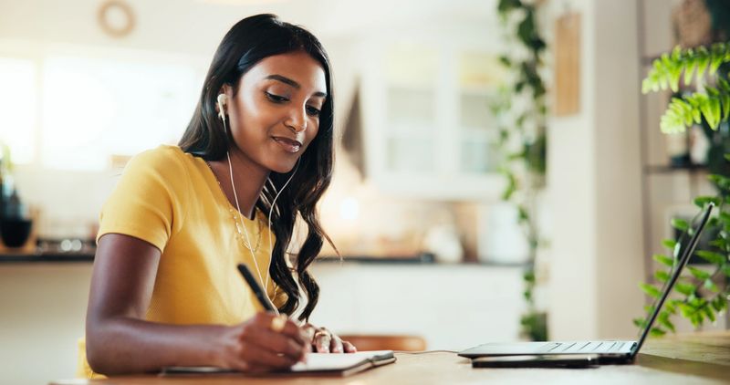 Laptop, woman and writing notes for elearning, webinar course or remote education in home. Computer, college student and notebook in virtual meeting, study and earphones for lesson in online class