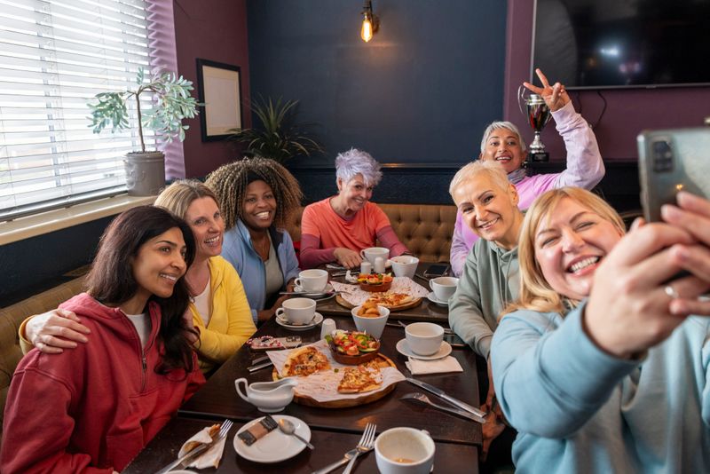 A wide shot of a cheerful group of diverse women enjoying brunch together, laughing and engaging in warm conversation. The cosy setting features a table filled with coffee, pizza, salads, and other delicious dishes. The women are all looking at a mobile phone as they are taking a group selfie. A genuine moment of friendship, connection, and positive energy.Videos are available similar to this scenario.