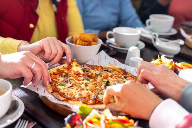 Close-up shot of a pizza and other foods on a table in a pub in Seghill, Northumberland. A group of women are having food and drink together after exercising. There are also various coffee cups and cutlery on the table. The women are taking a slice of pizza each.Videos are available similar to this scenario.