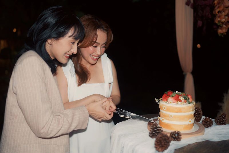 Newlywed LGBTQ couple is preparing to cut their wedding cake, looking into each other's eyes and smiling with happiness during this special evening moment.